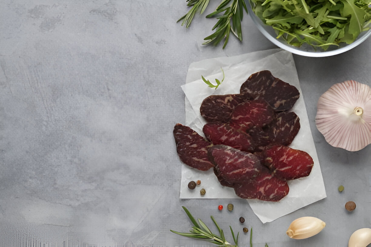 Slices of beef jerky arranged on parchment paper with fresh herbs and garlic on a gray background.