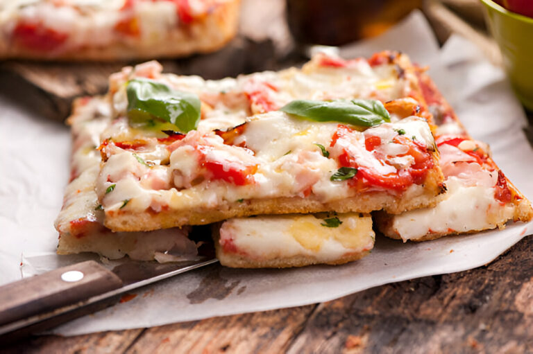A close-up of delicious rectangular pizza topped with melted cheese, red peppers, and fresh basil leaves on a rustic wooden surface.