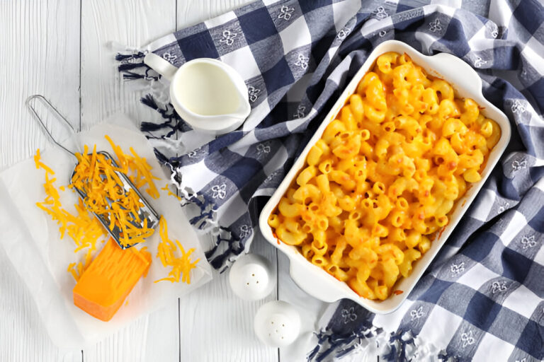 Baked macaroni and cheese served in a white dish with shredded cheddar cheese and milk on a blue and white checkered cloth.