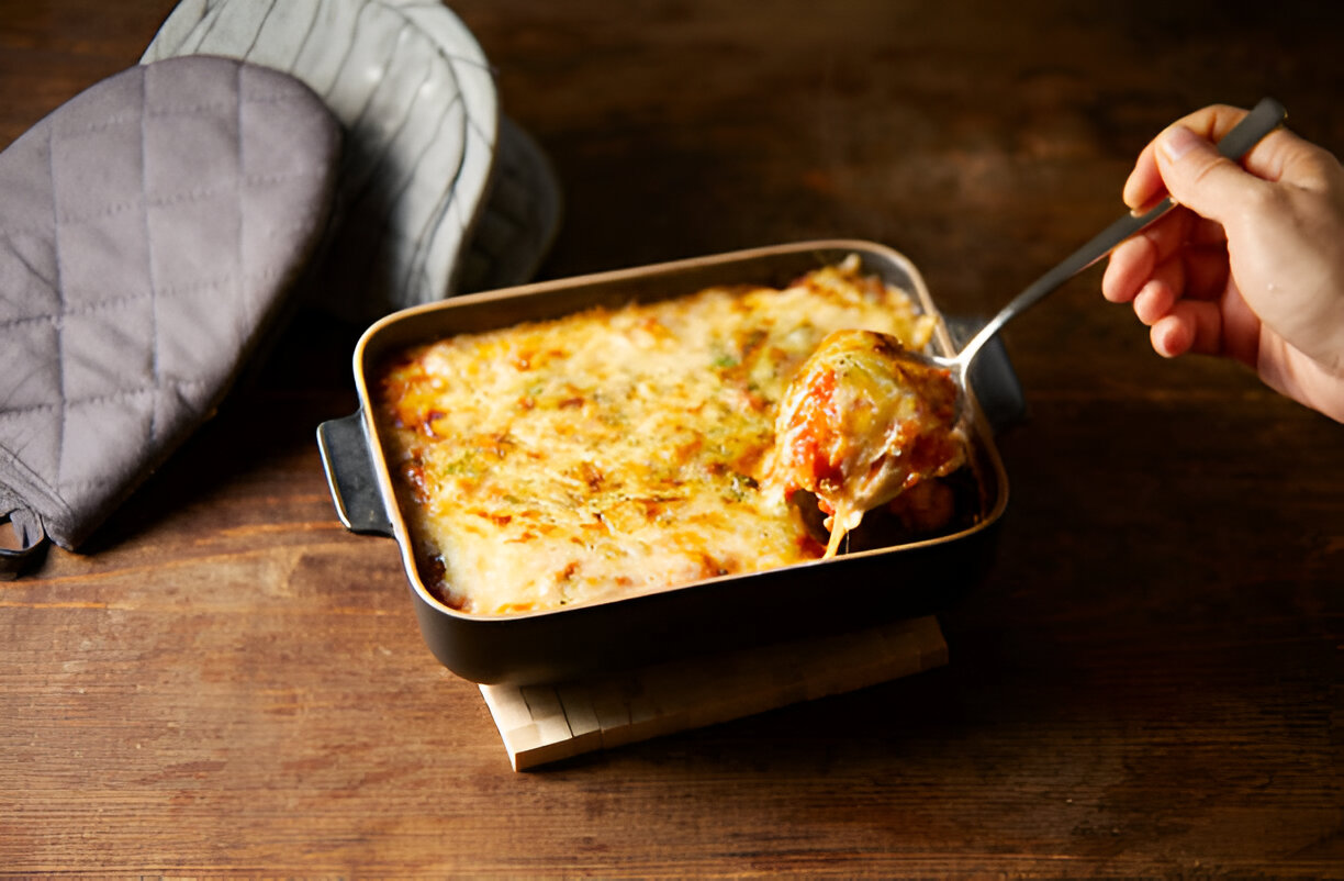 A hand holding a fork taking a portion of baked casserole from a black dish placed on a wooden table.