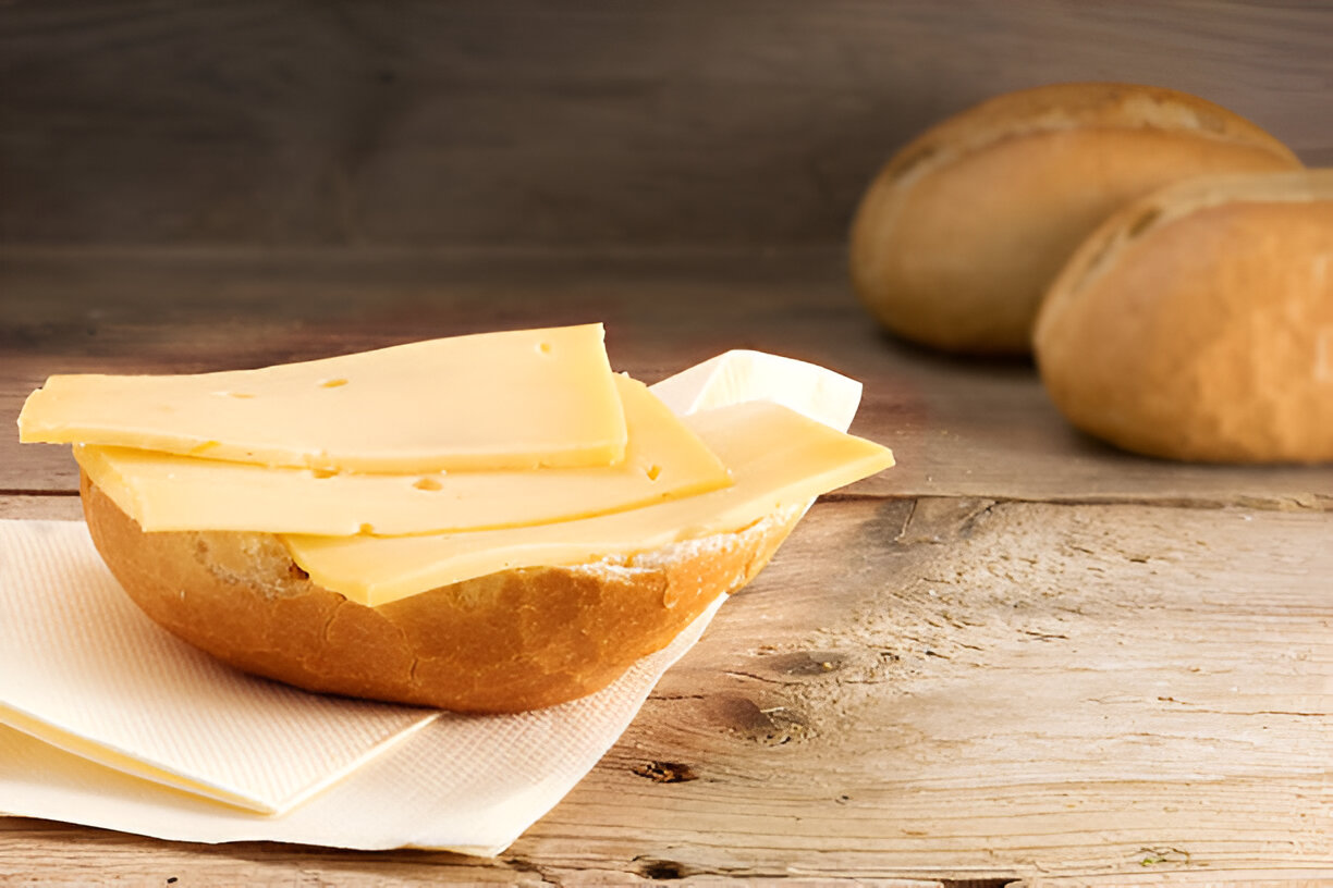 A soft bread roll topped with several layers of yellow cheese, placed on a napkin, with more bread rolls in the background.