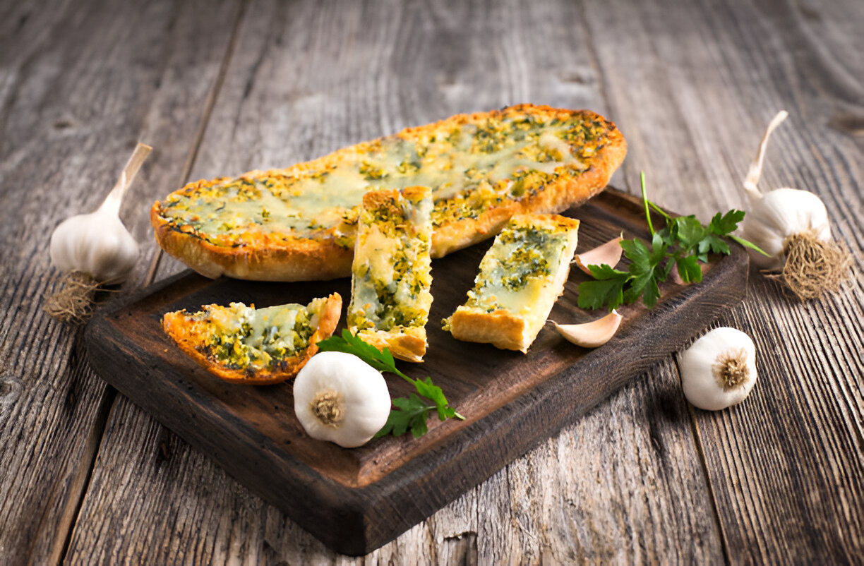 Freshly baked garlic bread with herbs on a wooden cutting board, surrounded by garlic cloves and parsley.