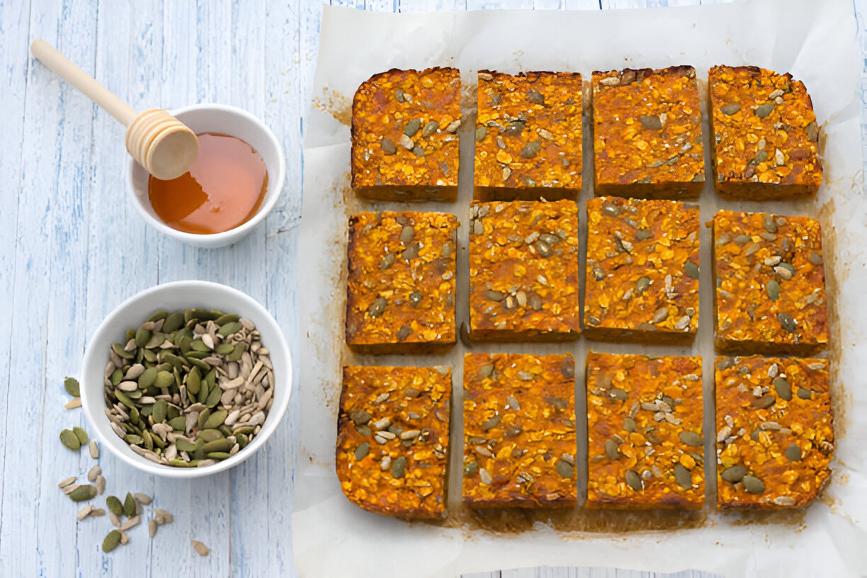 A freshly baked tray of pumpkin oat bars, cut into squares, displayed on parchment paper with bowls of pumpkin seeds and honey nearby.