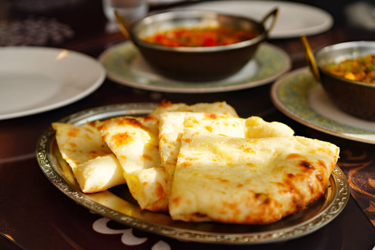 A plate of freshly baked naan bread with a golden brown crust, served on a silver platter, accompanied by Indian dishes in the background.