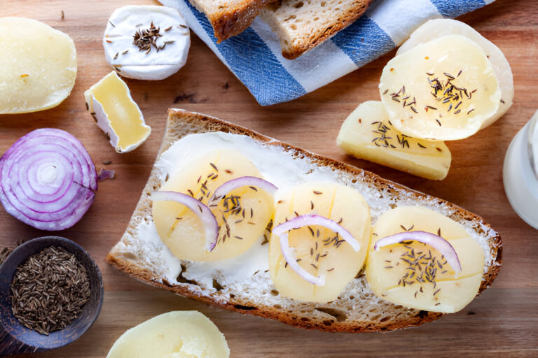 A rustic wooden board with slices of bread topped with cheese, potato, and onion, surrounded by additional cheese, red onion, and spices.