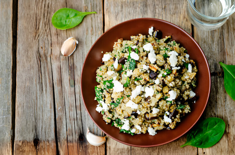 A bowl of quinoa salad topped with feta cheese, spinach, and garlic on a wooden table.