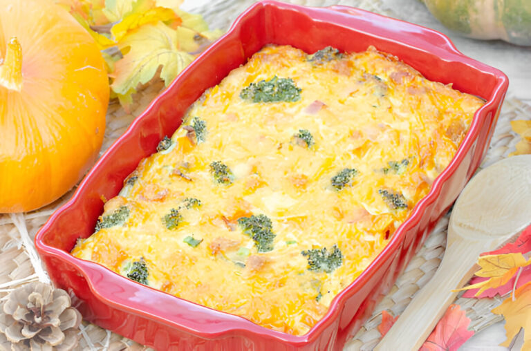 A baked dish with a golden, cheesy top and green broccoli pieces, placed in a red casserole dish, alongside a small pumpkin and autumn leaves.