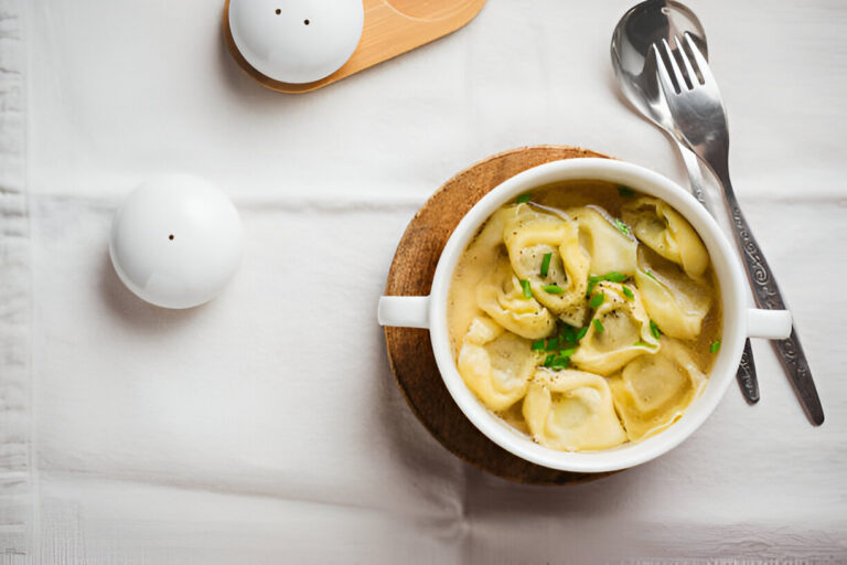A bowl of dumpling soup with fresh herbs, served with silver utensils on a white tablecloth.