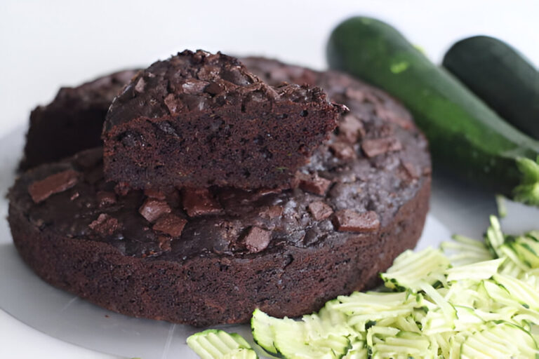 A chocolate zucchini cake topped with chocolate chunks, with sliced zucchini on the side.