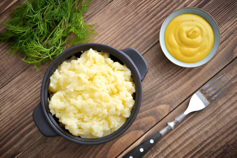 A bowl of creamy mashed potatoes served with a side of mustard and fresh herbs on a wooden surface.