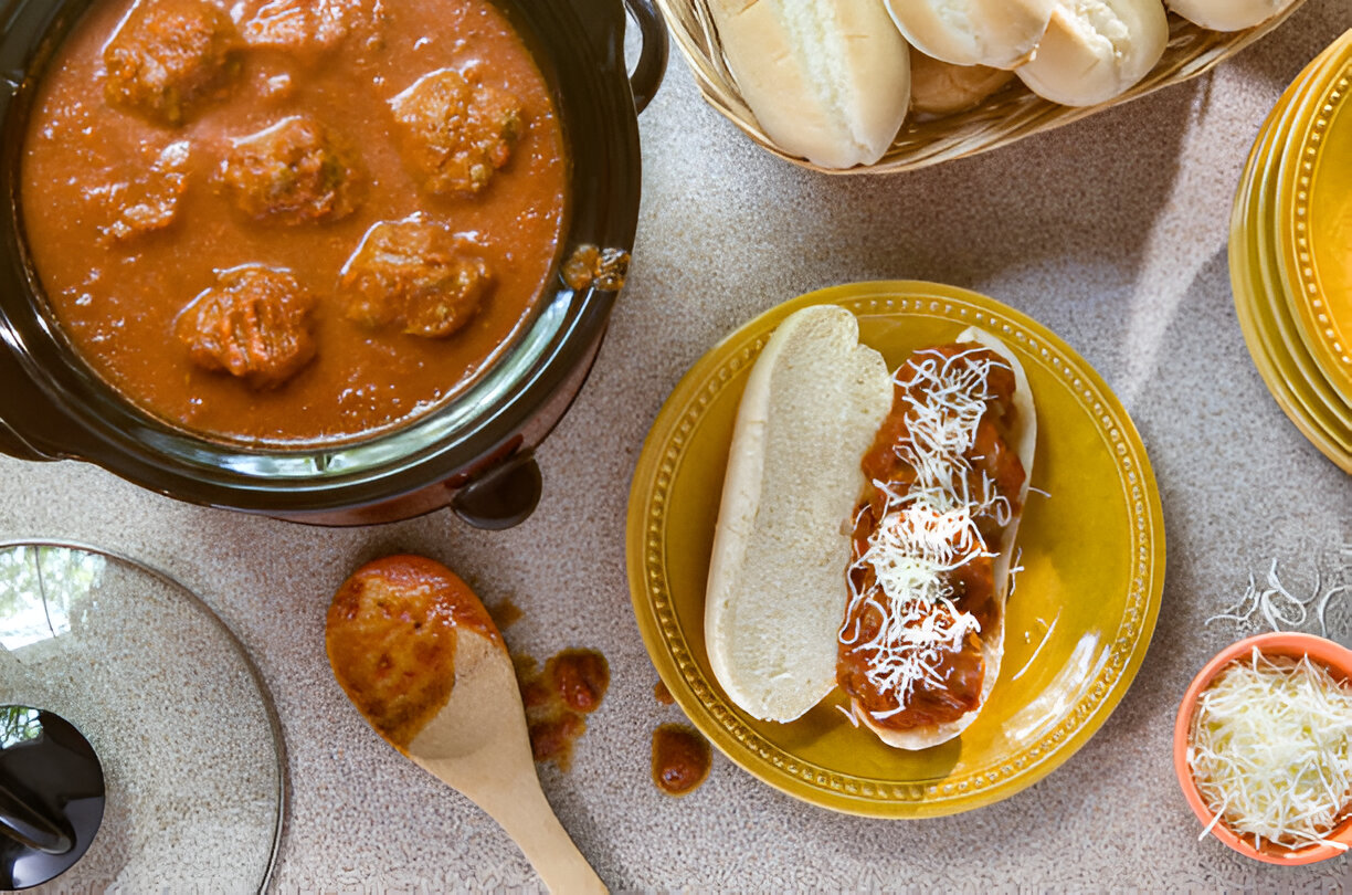 A plate of meatballs topped with marinara sauce and shredded cheese, alongside a soft bun and a bowl of shredded cheese.