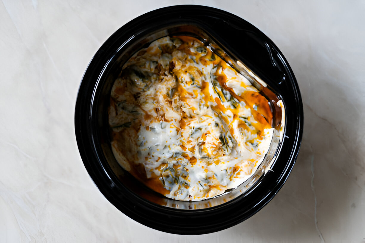 A close-up view of a slow cooker filled with creamy spinach and white sauce, topped with a drizzle of orange sauce.