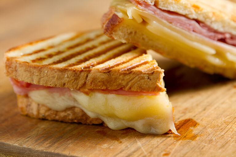 A close-up of a grilled sandwich with melted cheese and ham, sliced to show the filling, on a wooden cutting board.