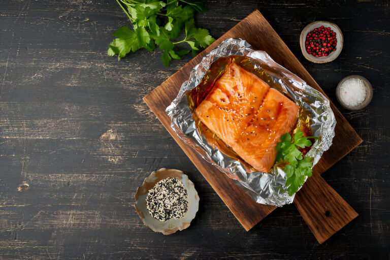 A piece of salmon fillet garnished with cilantro, placed in aluminum foil on a wooden cutting board, accompanied by sesame seeds, salt, and red spices in small bowls.