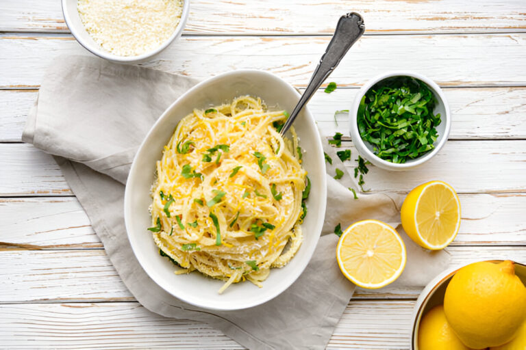 Bowl of pasta with herbs on a white wooden table, accompanied by lemons and grated cheese.