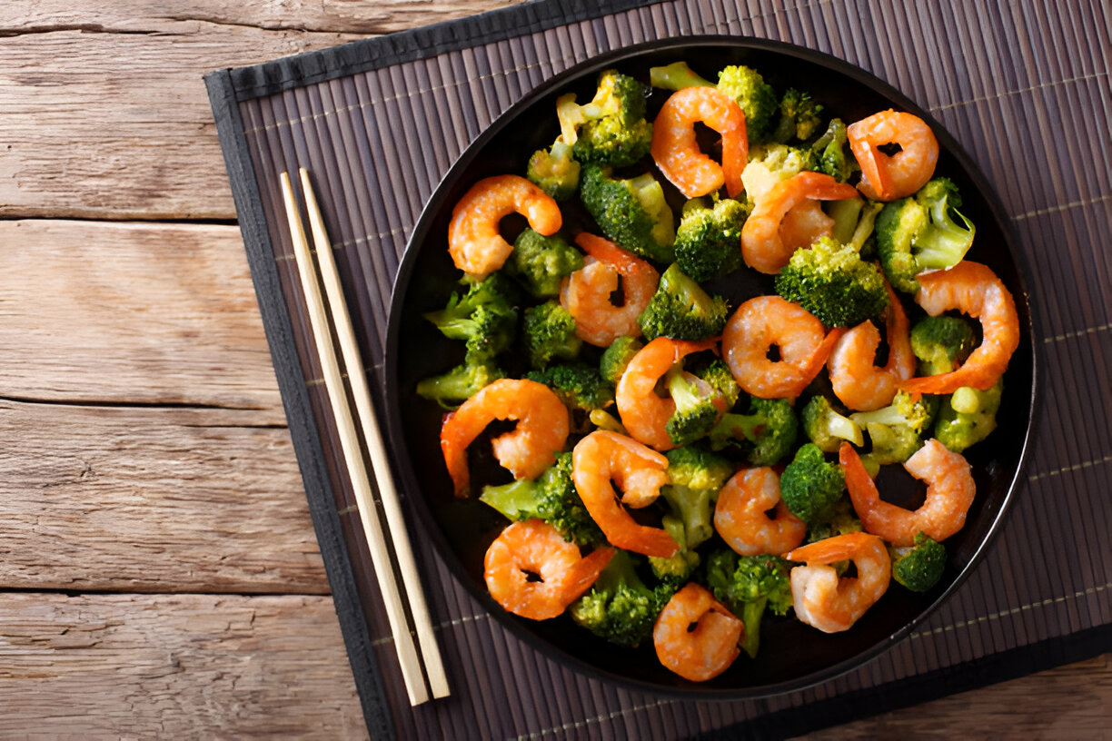 A close-up of a black plate filled with shrimp and broccoli, served with chopsticks on a bamboo mat.