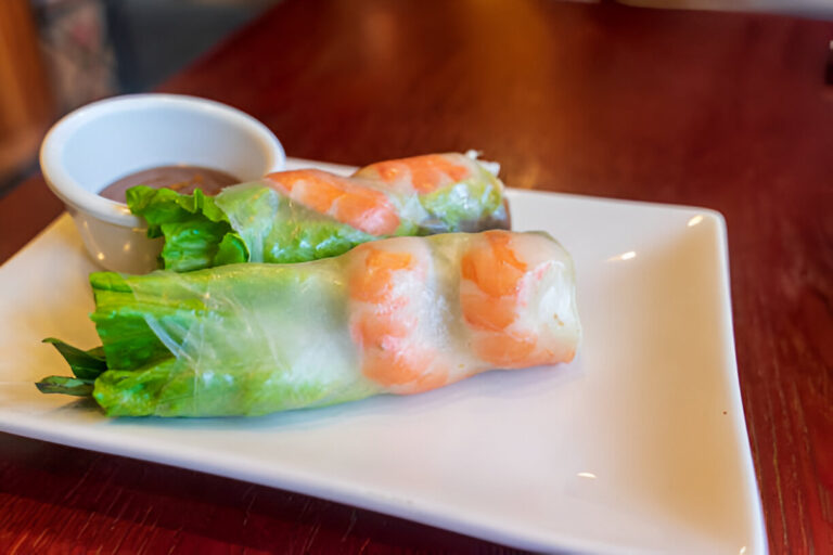 Two fresh spring rolls filled with shrimp and lettuce on a white plate, accompanied by a small bowl of dipping sauce.