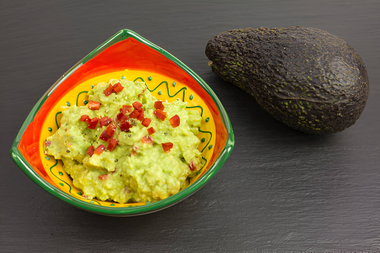 A vibrant green bowl filled with guacamole topped with diced red tomatoes, beside a whole avocado