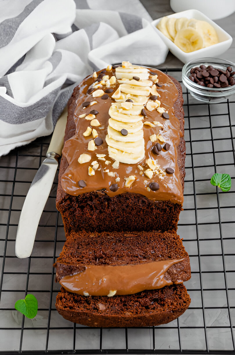 A sliced chocolate banana bread topped with banana slices, chocolate chips, and chopped nuts on a wire rack