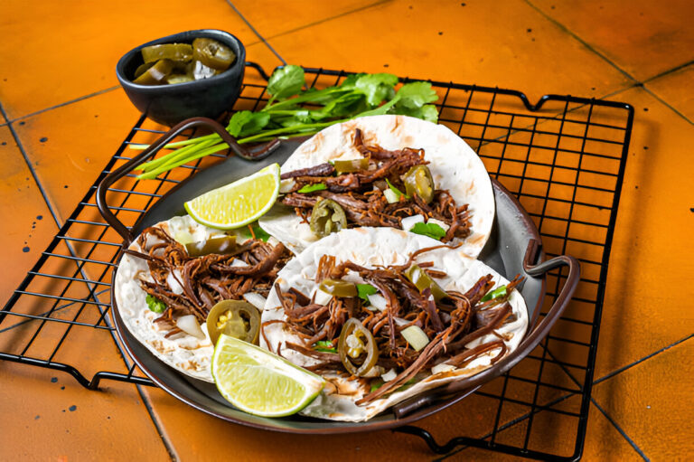 Three delicious tacos filled with shredded meat, garnished with jalapeños, onions, and lime wedges on a wire rack.
