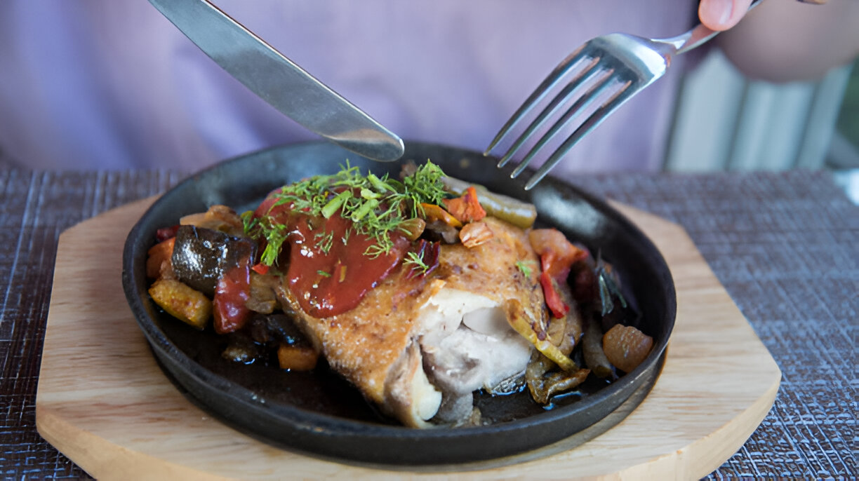 A plated chicken dish with vegetables on a hot stone plate, with a person using a knife and fork to cut it.