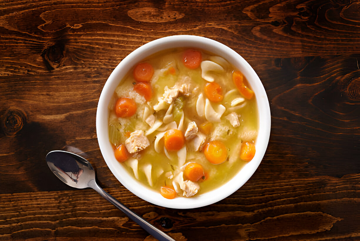 A bowl of chicken noodle soup with carrots and pasta on a wooden table.
