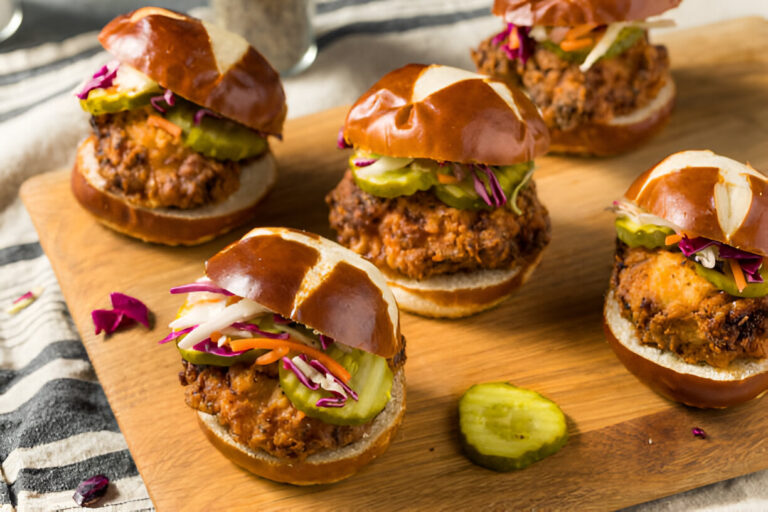 Five pretzel chicken sliders with pickles and slaw on a wooden cutting board.