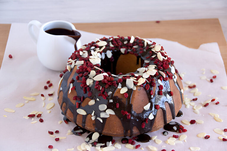A delicious bundt cake topped with chocolate glaze, dried raspberries, and sliced almonds, with a small pitcher of chocolate sauce beside it.