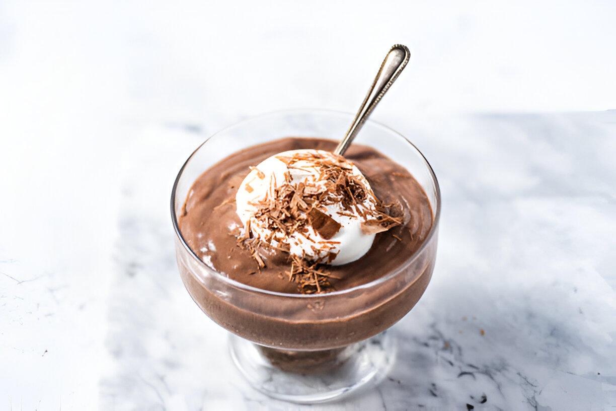 A delicious chocolate mousse topped with whipped cream and chocolate shavings in a clear glass dish with a spoon.