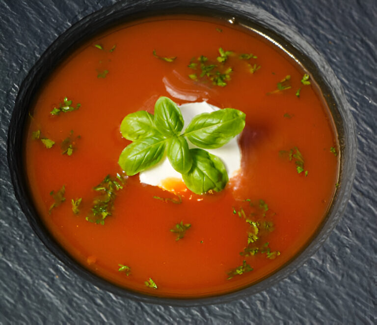 A bowl of tomato soup garnished with basil leaves and a dollop of cream, resting on a dark slate surface.