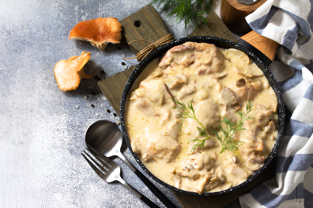 A delicious dish of creamy mushroom stew in a black skillet, placed on a wooden cutting board, accompanied by mushrooms and cutlery.
