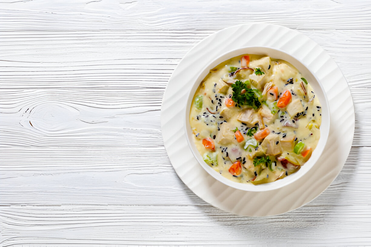 A bowl of creamy chicken soup garnished with herbs and vegetables, served on a textured white wooden table.