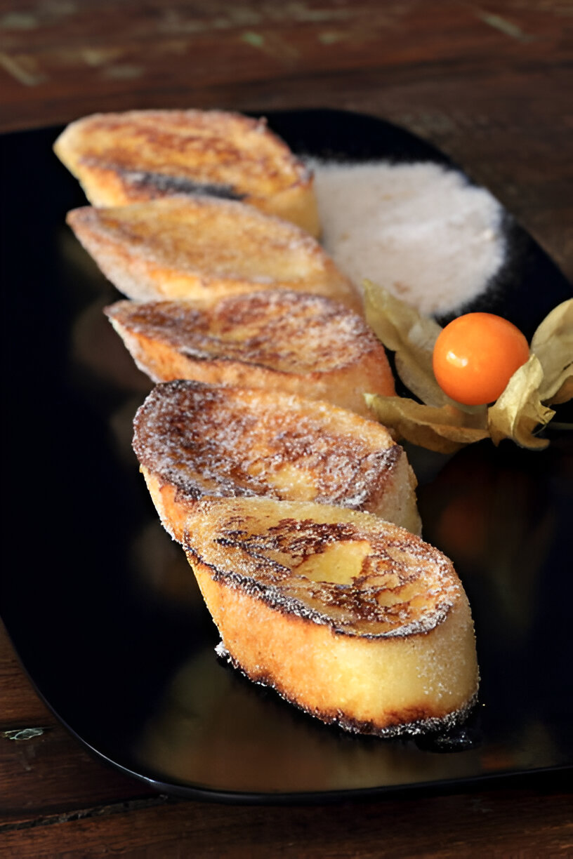 A platter of golden-brown fried bread slices sprinkled with sugar, accompanied by a small mound of sugar and a whole fruit.