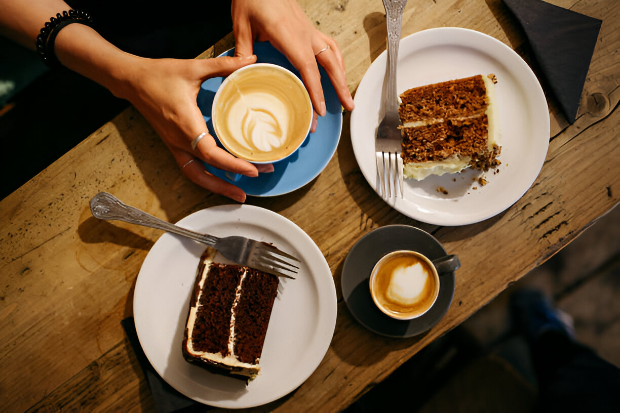 A hand holding a coffee cup beside two plates of cake on a wooden table.