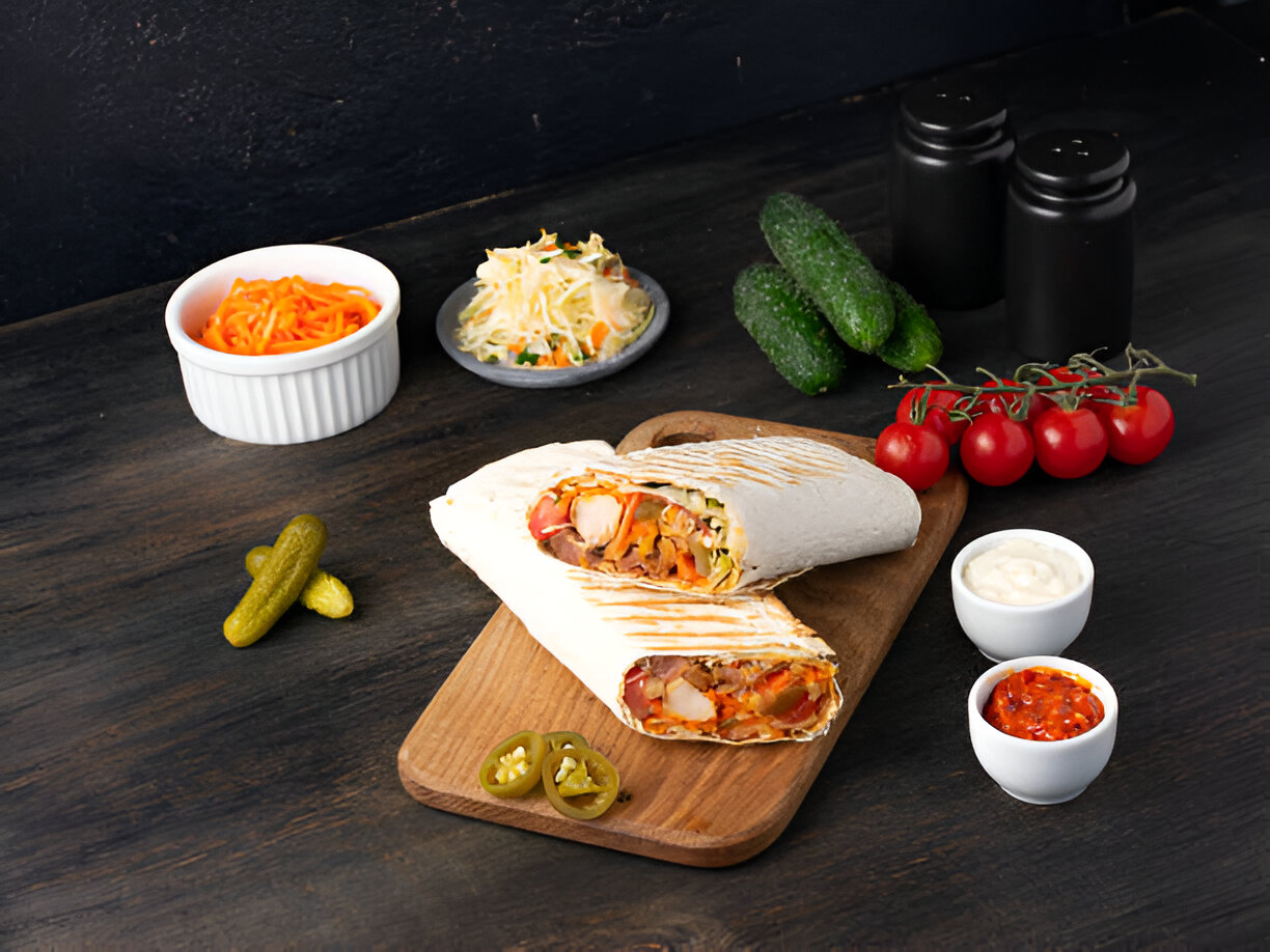 A delicious wrap with colorful vegetables, served on a wooden platter alongside fresh cucumbers, cherry tomatoes, pickles, and sauces.