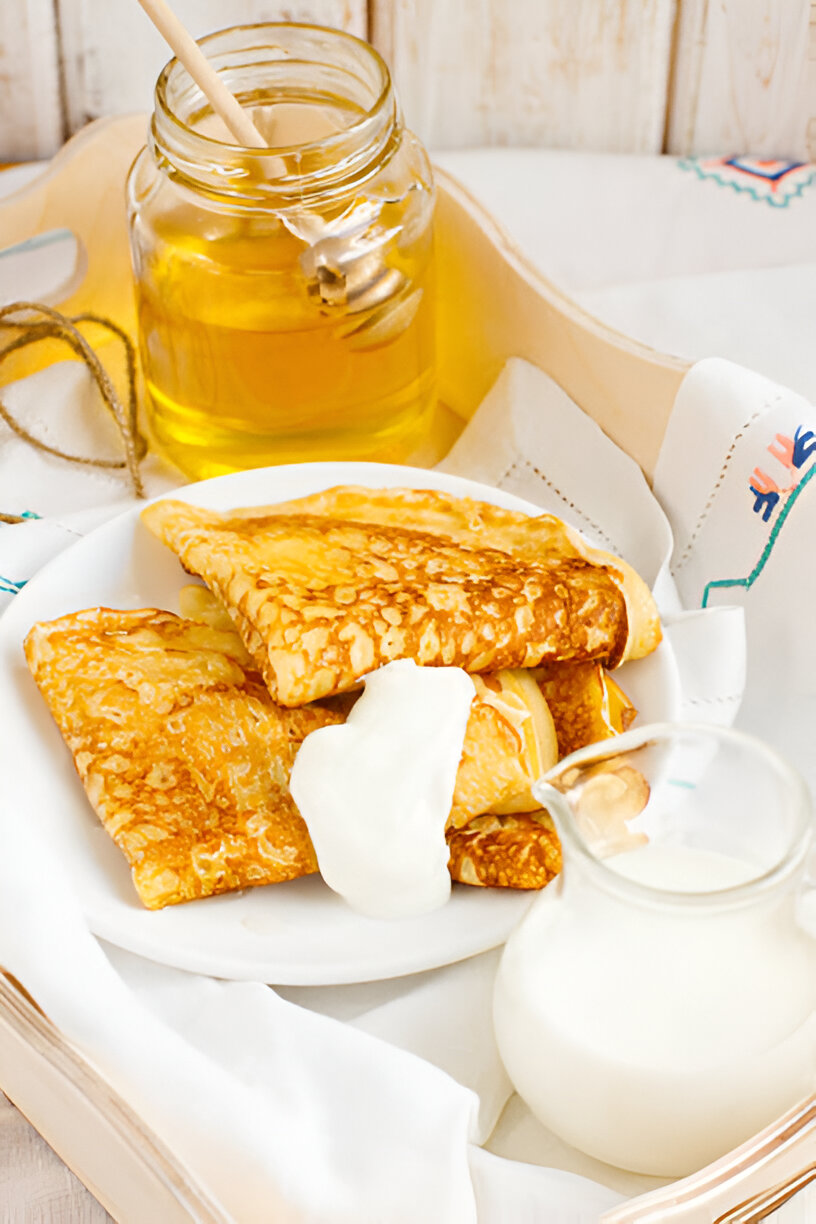 A plate of golden crepes topped with sour cream, accompanied by a jar of honey and a small jug of milk.