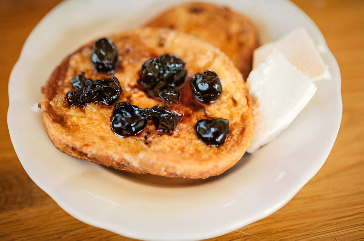 A close-up of a plate featuring two pieces of sweet fried bread topped with dark berry preserves and a small portion of white cheese.