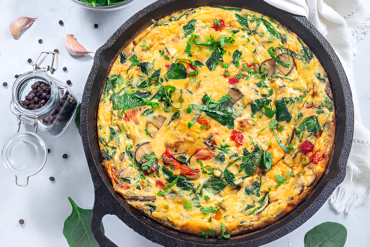 A freshly baked frittata loaded with spinach, tomatoes, and various herbs in a cast iron skillet on a white surface.