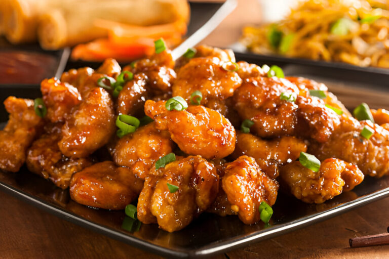 A delicious plate of sweet and sour chicken garnished with green onions, served with spring rolls in the background.