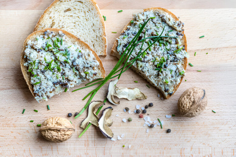 Slices of bread topped with a creamy spread, garnished with chives, accompanied by nuts and dried mushrooms on a wooden surface.