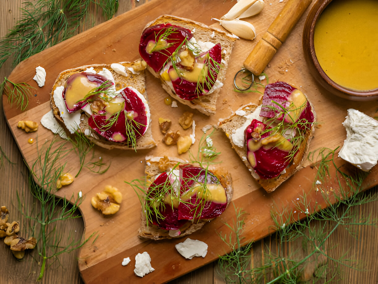 Sliced bread topped with beetroot, cheese, walnuts, and garnished with dill placed on a wooden cutting board