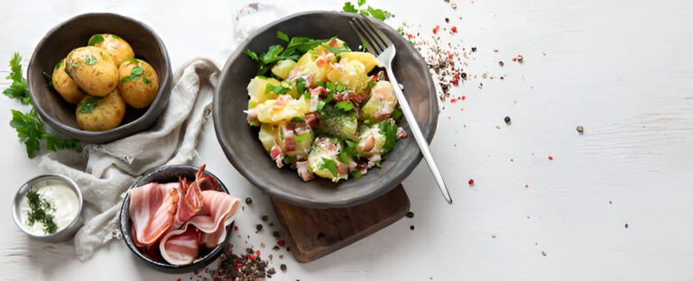 A bowl of colorful potato salad with diced vegetables and ham, accompanied by golden boiled potatoes and a bowl of sour cream on a white surface.