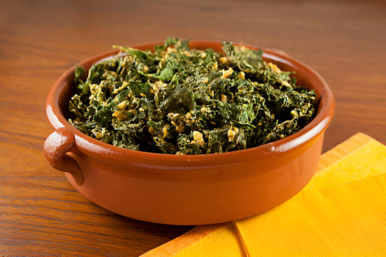 A brown ceramic bowl filled with cooked leafy greens on a wooden table with a yellow napkin beside it.