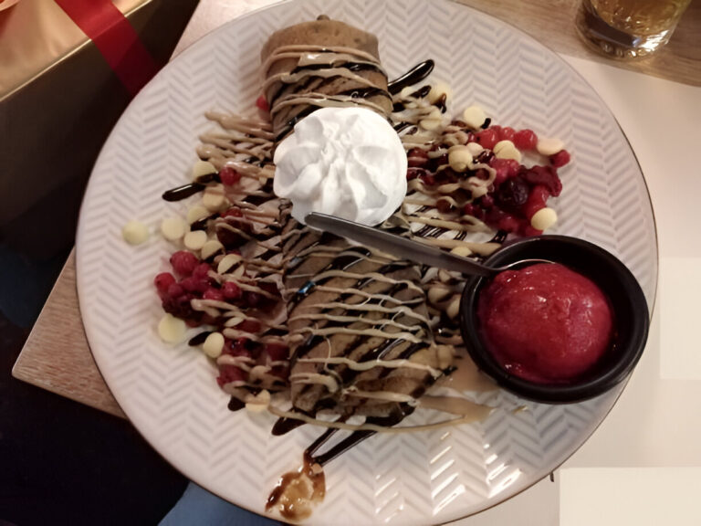 A plate of dessert featuring a crêpe topped with whipped cream, chocolate drizzle, and fresh berries, alongside a scoop of raspberry sorbet.