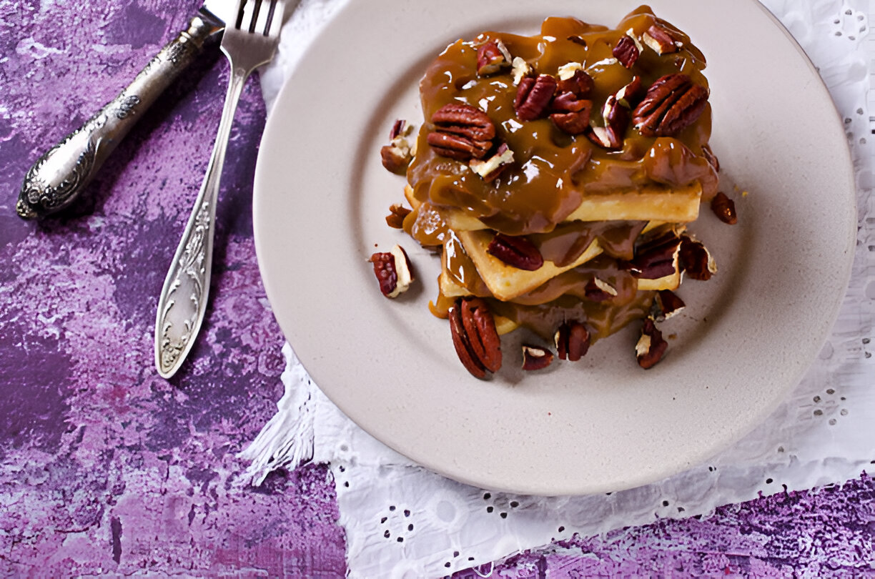 A plate of waffles topped with a caramel sauce and pecans, placed on a purple textured surface.