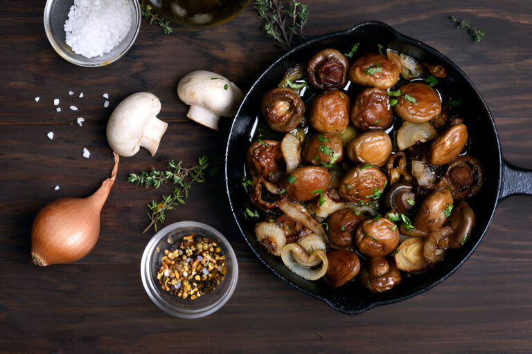 A cast iron skillet filled with sautéed mushrooms and onions, surrounded by fresh herbs, shallots, and seasoning.