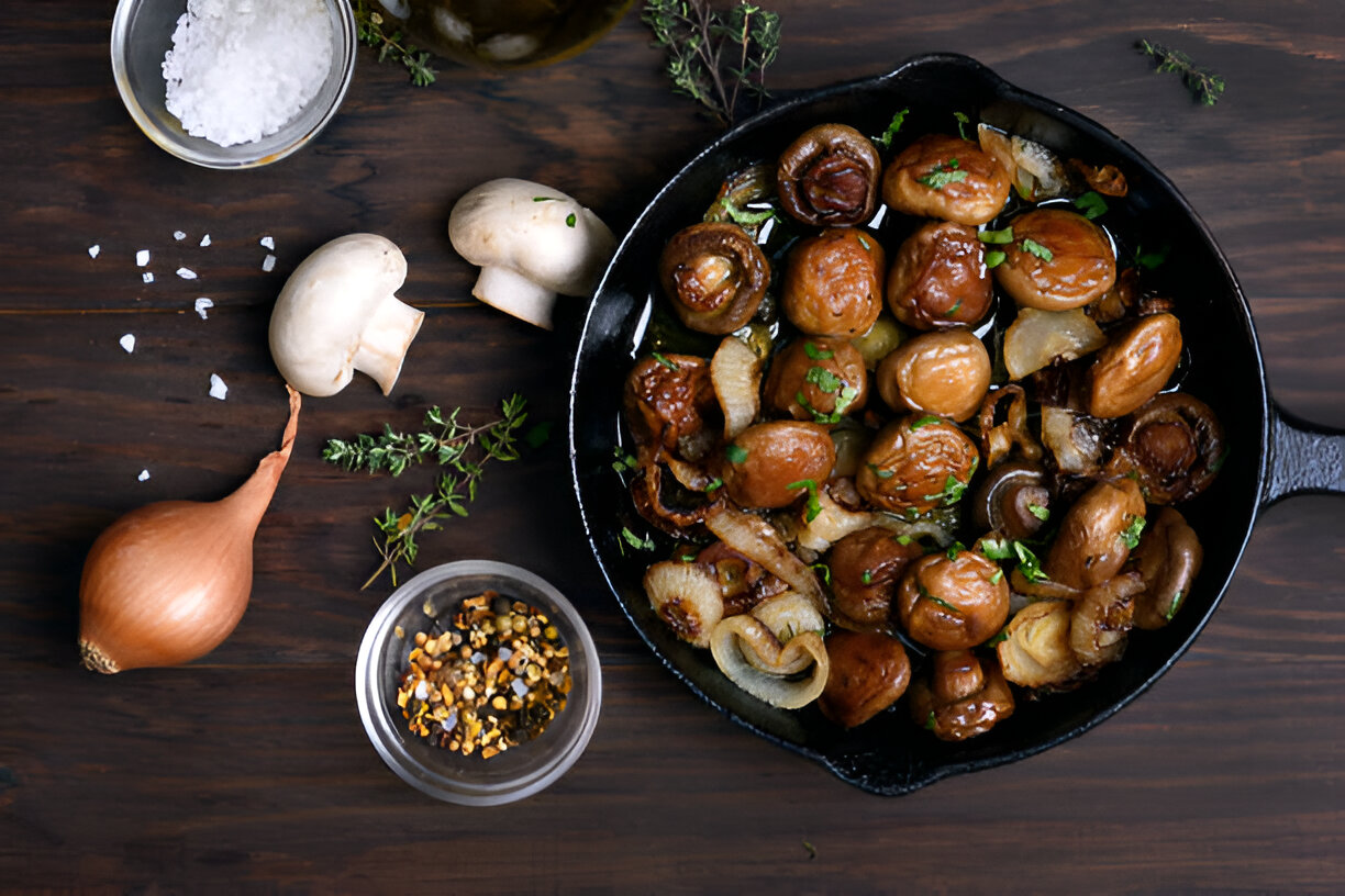 A cast iron skillet filled with sautéed mushrooms and onions, surrounded by fresh herbs, shallots, and seasoning.