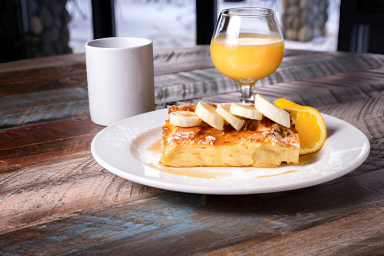 A plate of delicious French toast topped with banana slices and a slice of orange, accompanied by a cup of coffee and a glass of orange juice on a rustic wooden table.
