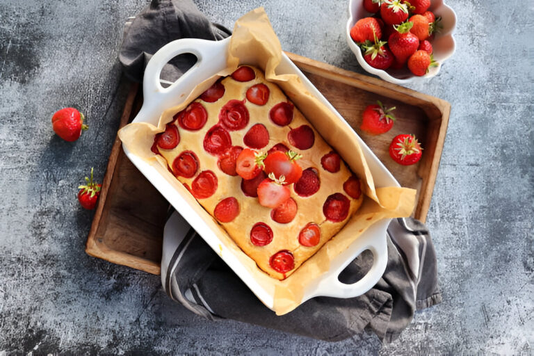 A freshly baked strawberry cake in a white baking dish, garnished with whole strawberries on top, placed on a wooden tray with a grey cloth.