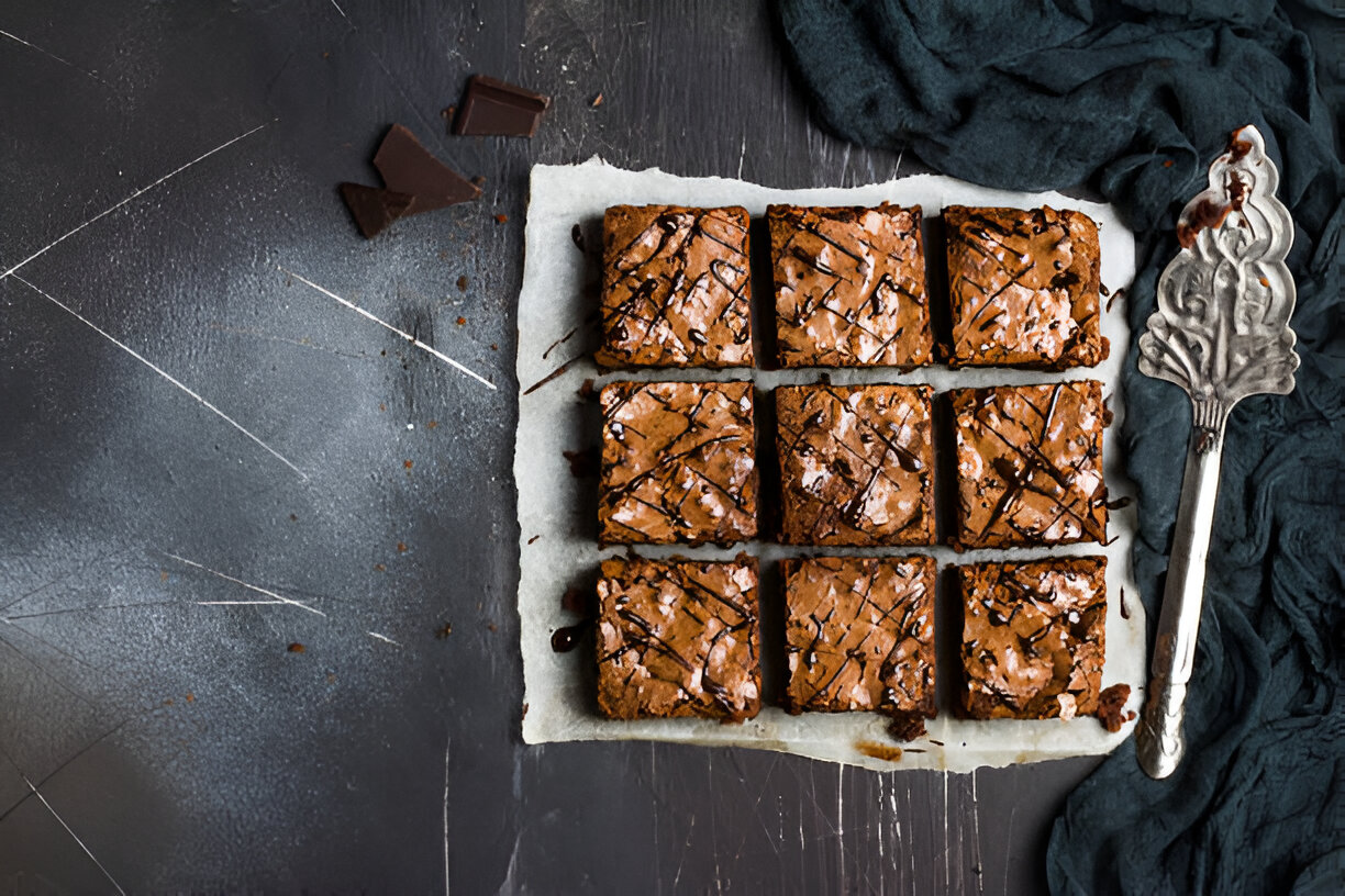 Delicious brownies arranged in a grid on parchment paper, drizzled with chocolate, next to a vintage serving spatula and a dark cloth.